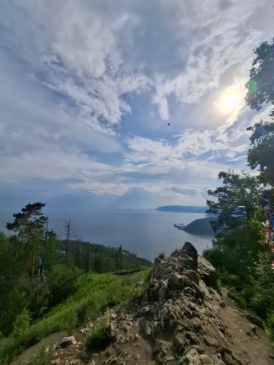 This is a landscape photograph taken from a high vantage point overlooking a body of water, likely a lake or river, in Nikola, Russia. The image is oriented vertically, with the sky and sun appearing on the left side and the land on the right.
The sky is partly cloudy, with bright sunlight breaking through from the upper left, casting a warm glow and creating lens flare. Wispy, elongated clouds stretch across the frame, suggesting a breezy atmosphere. A solitary bird is visible in flight against the sky. The water body occupies the central and right portions of the image, appearing calm and reflecting the light from the sky. In the distance, a distinct geological formation or island rises from the water.
The foreground on the right is dominated by a rocky, uneven terrain with patches of green grass and some sparse vegetation. Beyond this, a slope covered in lush green trees and undergrowth descends towards the water. A winding dirt path can be seen traversing the grassy slope. Some dead, bare trees are interspersed with the living foliage, adding a stark contrast to the vibrant greenery. At the very top of the image, a cluster of colorful, possibly prayer flags or decorations, are visible amongst the leaves of trees.
The overall impression is one of a natural, scenic vista on a sunny day, likely during the warmer months given the abundant greenery. The elevated perspective offers a panoramic view of the landscape, with a sense of openness and tranquility. No people or specific objects are clearly discernible, apart from the natural elements and the distant landmass. The time of day appears to be late morning or early afternoon, judging by the sun's position and intensity. There is no visible text in the image.