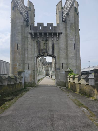The image captures a view of Conwy Castle in Llandudno Junction, United Kingdom. The scene is viewed through a stone archway of the castle. The archway leads to a paved walkway and a bridge stretching into the distance, passing through additional arches of the castle. The castle walls are constructed from large grey stones, showing signs of age and weathering, with green moss growing in places. There are crenellations on the towers. The paved walkway is made of a grey material, possibly asphalt or concrete, with patches of grass along the edges. A few people are walking on the bridge in the distance. A modern bridge is seen overhead, partially obscured by the castle walls. The sky above is pale blue with a few clouds.