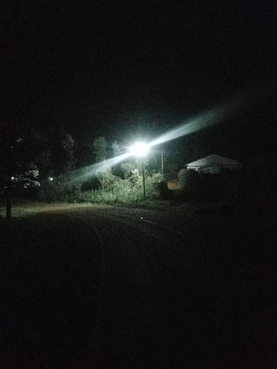 The image displays an outdoor night scene in Karuri, Kenya. A single, intensely bright street light, mounted on a utility pole, is centrally positioned, emitting a strong, diffused beam of light. Dense, dark green vegetation, consisting of bushes and trees, frames the illuminated area and extends into the background. In the upper right background, a building with a distinct light-colored, sloped roof is partially visible, largely obscured by the darkness and surrounding foliage. The foreground consists of a dark, unpaved or gravel path or road, exhibiting faint linear indentations or tracks. A small, light-colored object rests on the ground in the mid-right foreground. No individuals are present in the visible area.