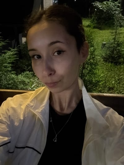 A close-up, waist-up, selfie-style photograph features a young woman sitting on a wooden bench outdoors at night. She has dark hair pulled back and is looking directly at the camera with a neutral expression. She is wearing a black t-shirt and a light-colored jacket with black trim. A silver necklace with a heart pendant is visible. The background consists of dark foliage and grass, with some artificial lighting illuminating the scene, suggesting an urban park or garden setting. The location is identified as Doma 1096 Km, Russia.