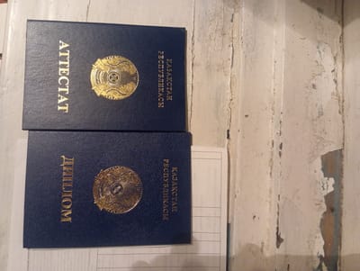 This media file features two dark blue diplomas with gold lettering and emblems, placed on a textured, light-colored surface. The diplomas are oriented vertically. The one on top has the text "ҚАЗАҚСТАН РЕСПУБЛИКАСЫ" on the left and "ДИПЛОМ" on the right, with a circular emblem in the center of the left side. The diploma below it has "ҚАЗАҚСТАН РЕСПУБЛИКАСЫ" on the left and "АТТЕСТАТ" on the right, also with a circular emblem on the left. The surface beneath the diplomas appears to be wood with a distressed, painted finish, showing signs of wear and discoloration. Faint lines of a document are visible under the top diploma. The overall scene suggests an indoor setting, possibly a desk or table, in a location within Kazakhstan, given the text on the diplomas. There is no indication of the time of day or weather conditions. The image is an object-focused still life, with the diplomas as the main subjects.