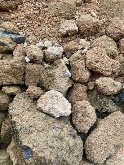 The image shows a pile of large, irregularly shaped rocks and clumps of compacted dirt. The rocks vary in size and color, ranging from light tan to dark brown and grey. Many of them have a granular texture, appearing to be composed of sand and small pebbles cemented together. Some rocks show patches of green algae or moss, suggesting they have been exposed to moisture. The ground beneath and around the rocks is also covered in loose dirt and smaller stones. The scene appears to be outdoors, possibly in a construction site or an uncultivated area. There are no people or animals visible in the image. The lighting suggests it is daytime, with diffused light. There is no specific context provided about the location within Bulamare, Nigeria.