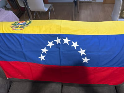 The image shows a flag of Venezuela laid out on a piece of furniture, possibly a sofa or couch, in an indoor setting. The flag is composed of three horizontal bands of equal width: yellow on top, blue in the middle, and red on the bottom. In the blue band, there is an arc of eight white five-pointed stars. In the upper left corner of the yellow band, the Venezuelan coat of arms is depicted. The coat of arms features a shield divided into three sections, with a horse in the central section, and wheat and a sword in the side sections. The background of the image shows furniture, including chairs with white upholstery and metallic legs, and wooden flooring, suggesting a domestic or communal space. The lighting appears to be artificial. The provided metadata indicates the image was taken in Getafe, Spain.