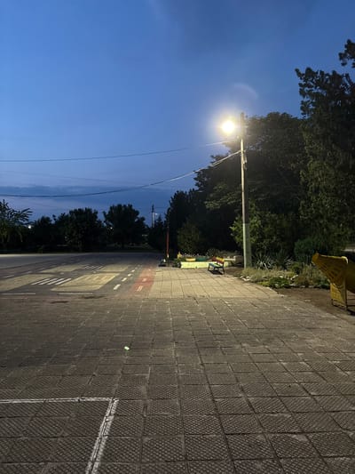 The image depicts an outdoor scene at dusk or early night in Saedinenie, Bulgaria. The sky is a deep blue with faint cloud formations, crossed by multiple dark power lines. A single, brightly illuminated street light on a tall utility pole casts a warm glow, primarily on the paved ground and nearby vegetation.

The foreground and midground feature a patterned, dark grey paved area comprised of square, textured tiles. This pavement is marked with white and a section of red paint, forming miniature road lines, including what appears to be a crosswalk and lane divisions, suggesting a children's traffic park or training area. To the left, the paved area expands into what resembles a miniature road or open space, bordered by dark, indistinct trees in the distance.

On the right side of the image, dense foliage and trees, mostly dark due to the low light, form a natural boundary. Positioned near the street light and against the vegetation is a wooden bench painted with horizontal stripes in yellow, red, blue, and green. Adjacent to the bench are small planted garden beds with some visible flowers. Further to the right, a large, yellow, possibly metal structure, partially visible, stands near the edge of the scene; it appears to be an outdoor fixture like a stylized trash bin or a play element. No people are visible in the scene. A small, bright green object lies on the paved ground in the foreground.