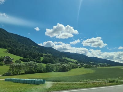 A sunny day over a lush green landscape in Scheifling, Austria, features rolling hills dotted with trees and buildings, beneath a bright blue sky filled with white, fluffy clouds. In the foreground, a row of teal-wrapped hay bales sits on a green field. A paved road with white lines runs along the bottom edge of the image. The backdrop is a majestic mountain range covered in dense green forest, with scattered houses and farm buildings nestled on its slopes. The sun's rays create a bright flare in the upper right corner of the sky.