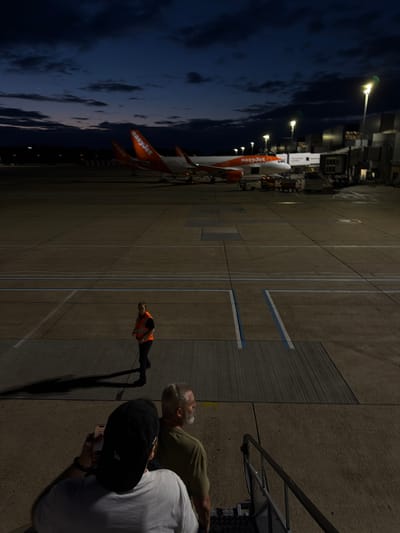 The image depicts an airport tarmac at dusk, likely at London Gatwick Airport (LGW) in Crawley, United Kingdom. Two orange and white EasyJet airplanes are parked at gates. In the foreground, two individuals are visible from behind, one wearing a black baseball cap and holding a smartphone. Further back, an airport worker in an orange high-visibility vest is walking across the tarmac. The scene is illuminated by artificial lights and the fading daylight, with a dark, cloudy sky overhead. The airport environment includes jet bridges and service vehicles.