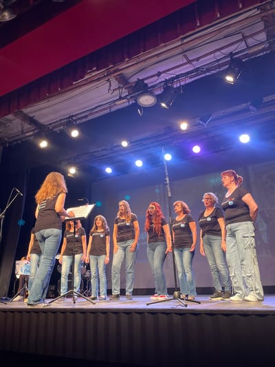 A group of eight women wearing black t-shirts with a white logo and blue jeans are on a stage, performing. The woman on the far left, facing away from the camera, appears to be directing the group. The other seven women are facing the audience, singing. They are standing in two rows, with four women in the back and three in the front. The stage has a dark backdrop and is lit with bright stage lights. There are microphones and a music stand on stage. The overall scene suggests a musical performance, possibly a choir or a vocal group. The presence of a sign with "AUTUMN CONCERT" written on it indicates this is part of a concert event. This concert is part of the "FESTES DE LA MARE DE DÉU DE LA MERCÈ" celebration in Arenys de Mar, Spain.