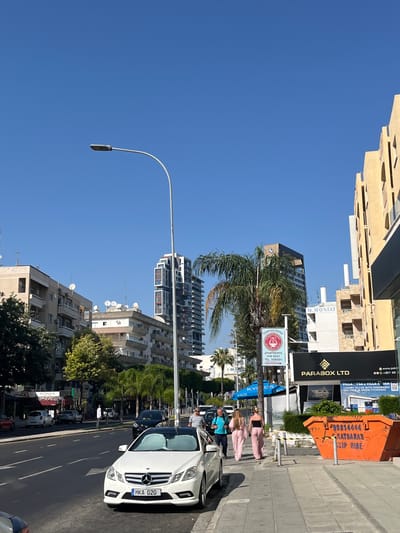 The image depicts a street scene in Germasogeia Municipality, Cyprus, under a clear blue sky. In the foreground, a white Mercedes-Benz sedan with license plate "MKA 020" is parked on the right side of the road. To its right, a bright orange skip bin labeled "99814444 KATSARAS SKIP HIRE" is visible. Further down the sidewalk, a group of people are walking away from the camera: two women in matching pink outfits, and a man in a blue t-shirt and dark shorts. Behind them, a man in a striped shirt and straw hat is walking towards the camera.
The street is lined with buildings of various architectural styles. On the left, there are apartment buildings with balconies and satellite dishes. In the middle ground, a tall, modern skyscraper with a distinctive geometric design dominates the skyline, with palm trees in front of it. To the right, another building with a beige facade and arched windows is prominent. Signs visible include "TERPSICHORE APARTMENTS FOR RENT", "M. MONIAT", "PARABOX LTD", and "KYKOS". The road has multiple lanes, and several cars are parked or driving on it. The environment appears to be an urban setting with a mix of residential and commercial structures.