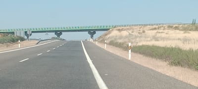The image depicts a view from a moving vehicle on a highway near Miajadas, Spain. The highway is asphalt with white dashed and solid lines indicating lanes. A green overpass with concrete supports spans across the highway in the distance. The sides of the highway are bordered by dry, grassy embankments and sparse vegetation. Several white posts with orange reflective markers are visible along the roadside, indicating the edge of the road. The sky is clear and blue, suggesting a sunny day. The scene is devoid of any people or other vehicles.