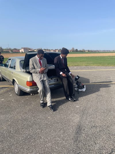 The scene depicts two Black males in suits positioned on a paved asphalt surface in Reventin-Vaugris, France, under a clear blue sky. A light green/gold Mercedes-Benz sedan with an open trunk is central. The male on the left, wearing a light gray suit and red tie, leans against the car's rear, looking down at a smartphone. The male on the right, in a dark suit, red tie, and glasses, sits on the open trunk's edge, looking at a professional video camera and monitor assembly on a small tripod placed on the ground beside him. A third male, partially visible on the far left, wears a dark shirt with white graphics. The car displays a French license plate: B-203-GC. The background features expansive green and fallow fields, leading to a distant cluster of buildings and trees.