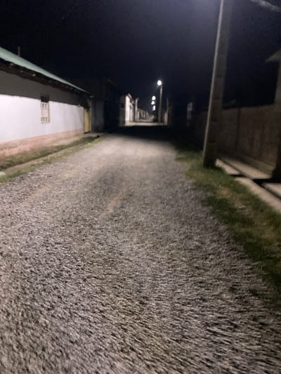 A low-angle, first-person perspective shot looking down a dark, gravel road at night. On the left, a white building with a green roof and a window with bars is visible. On the right, a tall, light-colored pole stands near a darker structure with a concrete base. Several streetlights are visible further down the road, casting light on the gravel and illuminating the night sky. The scene appears to be in a rural or suburban area, consistent with a location in Боғистон, Tajikistan.