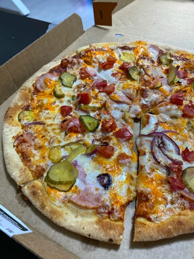 A close-up, overhead shot shows a pizza with various toppings, including pickles, tomatoes, red onions, and ham, resting in a cardboard pizza box. The pizza is sliced into pieces, with some pieces overlapping. The crust is golden brown and appears to be thick. The pizza is well-lit, and the colors of the toppings are vibrant. The background is blurred, but it appears to be an indoor setting. There is a small brown cardboard flap with the number "2626" printed in black on it, affixed to the inside of the box. No people or animals are visible. The scene depicts a pizza ready to be eaten.
