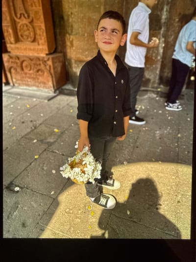 A young boy, likely between 8 and 10 years old, stands in what appears to be an outdoor courtyard with stone paving and scattered petals. He is wearing a black collared shirt with gold buttons and black pants. On his feet, he has black and white sneakers. In his right hand, he holds a small, woven basket filled with white flowers. He is looking upwards and to his left, with a slight smile on his face.

Behind him, two other individuals are partially visible. The person directly behind him is wearing a white polo shirt and dark pants. Further behind that, another person is wearing a light blue shirt and dark pants.

To the left of the boy, there are large, carved stone structures, possibly khachkars (Armenian cross-stones), with intricate designs. The ground is paved with cobblestones and is partially covered with fallen white and yellow petals, suggesting a recent event or ceremony. The lighting suggests it is daytime, with a strong shadow of the boy cast on the ground to his right. The overall scene seems to be set in a historical or religious site, consistent with the presence of khachkars. This location is identified as the courtyard of the Saint Gregory Cathedral in Artik, Armenia.