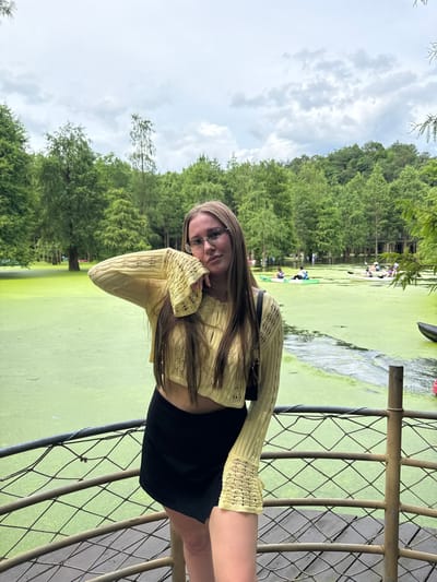 A young woman with long blonde hair and glasses, wearing a yellow cropped sweater and a black skirt, stands on a wooden platform with a metal railing in front of a murky green body of water. The water is covered in a thick layer of algae, and several people in kayaks can be seen in the background. The scene is set amidst a dense forest of trees, with a cloudy sky overhead. The location appears to be within the city of Lin'an District, China.