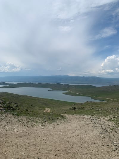 The image depicts a panoramic view of a vast body of water, likely a lake, bordered by rolling green hills under a cloudy sky. In the foreground, a dirt path winds down a rocky, grassy incline. The middle ground features the expansive water, with several peninsulas and islands jutting out into it. A small cluster of buildings is visible on one of the peninsulas, and a dirt road is seen meandering through the landscape. In the distance, a mountain range is faintly visible through a haze, with snow-capped peaks suggested on the horizon. The sky is a mix of grey clouds and patches of blue, with some white, puffy clouds scattered throughout. The overall impression is a serene, natural landscape. The location is the vicinity of Khuzhir, Russia, on Olkhon Island, which is the largest island in Lake Baikal.