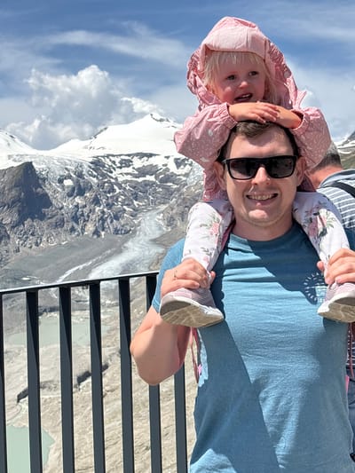 A man wearing sunglasses and a blue t-shirt carries a young girl on his shoulders. The girl, wearing a pink hooded jacket, rests her hands on the man's head. In the background, snow-capped mountains and a glacier are visible under a cloudy sky. A black metal fence runs across the foreground. The scene is set in Heiligenblut am Großglockner, Austria, known for its mountainous terrain.