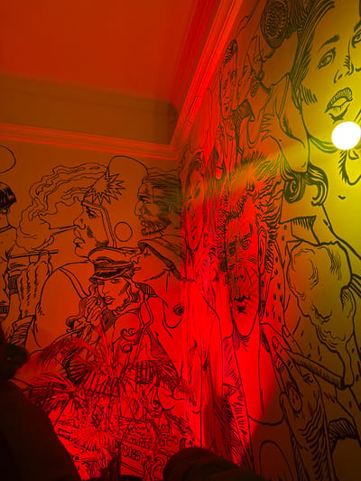 The image depicts a room in ქვედა სამება, Georgia, featuring walls and a ceiling covered in a black line art mural on a yellow background. The mural depicts a variety of figures, faces, and scenes. The room is illuminated by two light sources: a round, white overhead light and a red light emanating from the lower right corner. The red light casts shadows and a red hue over a section of the mural and a cluster of thin, linear objects, possibly plants. The left side of the room shows a doorway.