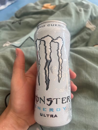 A hand is holding a white can of Monster Energy Ultra drink. The can has the Monster Energy logo and "ZERO CUKRU" written on it. The background is a green-blue patterned blanket. The scene appears to be indoors. The location of Szczebrzeszyn, Poland is not depicted or suggested by the image.