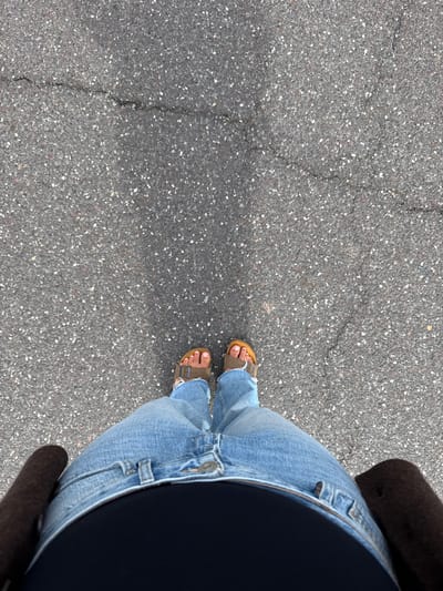 An overhead view depicts one individual standing on a dark gray asphalt surface. The person is visible from the upper thighs down, wearing light blue denim jeans with minor fraying at the cuffs and a dark, possibly black, shirt. On the individual's feet are open-toe sandals with dark olive green straps and tan soles, revealing bare feet with reddish-orange painted toenails. A dark brown object, possibly a sleeve or bag, is partially visible on the far left. The asphalt ground exhibits a textured appearance with embedded lighter aggregates and multiple fine, irregular cracks. Two distinct shadows, cast by the person's legs and feet, are visible on the asphalt, extending from the lower center towards the upper center of the frame. The image does not contain visual data to confirm the specified location of West Windsor, United States.
