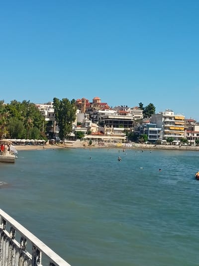 The image shows a view of a beach and town in the Municipal Unit of Anthidon, Greece. In the foreground, there is a body of water with several people swimming. The water is a greenish-blue color. In the middle ground, there is a sandy beach with rows of lounge chairs and umbrellas. Behind the beach, there are buildings, some of which appear to be hotels or apartments. There are also trees and other vegetation. The buildings are a mix of white and pastel colors, and many have balconies. The sky is clear and blue. On the left side of the image, there is a white railing. The overall scene suggests a sunny day at a coastal resort.