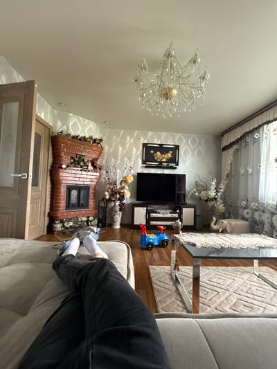 This image depicts an indoor scene, likely a living room, in what appears to be a home in Beryozovka, Russia. The room is furnished with a sofa, a coffee table with a glass top, a television set on a cabinet, and a decorative brick fireplace. A large, ornate chandelier hangs from the ceiling. The decor includes a framed artwork with gold leaf and a vase with artificial flowers. On the floor, a child's blue toy car is visible. The room is lit by artificial light, suggesting it is either evening or a dimly lit interior. The overall atmosphere appears relaxed and domestic, with a person's legs and feet visible on the sofa, wearing black pants and white socks. The image is oriented vertically, with the ceiling at the bottom and the floor at the top. There is no visible text in the image.