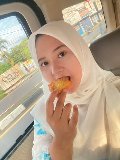A woman in a white hijab sits in a car and eats a fried pastry. Her head is tilted to the side, and she looks at the camera with dark eyes. Her right hand holds the pastry to her mouth. Outside the car window, a street with buildings and foliage is visible. The car's interior includes a headrest and a light-colored dashboard.