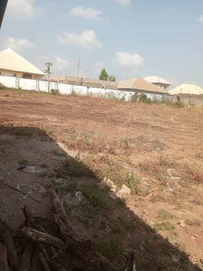 A residential neighborhood displays a mix of architectural styles behind a white perimeter wall. The ground is barren and brown, showing sparse vegetation. Shadows cast by the unseen structure where the photo was taken intersect with scattered debris, including wood and plastic. The sky is light blue with some clouds.