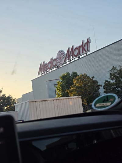 The image shows the facade of a MediaMarkt electronics store in Bischofsheim, Germany. The store has a large, white, corrugated metal exterior. A prominent red and white sign with the MediaMarkt logo is visible across the top of the building. The sky is a pale blue, suggesting it is either early morning or late afternoon. Green trees are visible on the right side of the building. In the foreground, the dashboard of a car is visible, with a sticker on the windshield or dashboard. The scene appears to be captured from inside a vehicle, looking out towards the store.