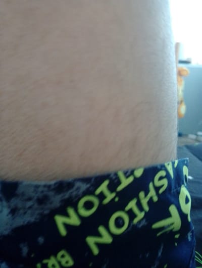 The image shows a close-up of a person's arm and a dark blue fabric with lime green lettering. The lettering appears to be a repeating pattern that includes the words "FASHION" and "ART". The fabric is wrinkled, suggesting it is clothing or a textile item. The person's arm is lightly haired and appears to be pale. In the background, out of focus, a blurred orange object and a light-colored wall or surface are visible. There are no discernible features of Krasnovishersk, Russia in this image.