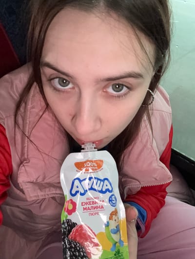 A young woman with brown hair and light eyes is drinking from a white and blue squeezable fruit puree pouch. The pouch features images of raspberries and blackberries. She wears a pink and red puffy jacket and what appears to be a light pink shirt. The background is blurry, suggesting an indoor environment. It is not possible to determine the location in Lunezhki, Russia.