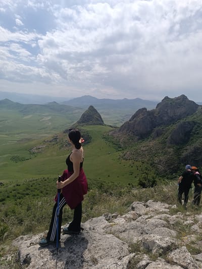 The image depicts a landscape with a woman standing on a rocky outcrop. The location is Hüseynbəyli, Azerbaijan. The woman is facing left, looking out over a broad, green valley dotted with hills and mountains in the distance. She is wearing a black tank top, black leggings with blue stripes, hiking shoes, and has a maroon jacket tied around her waist. She is holding two hiking poles.

In the background, to the right of the woman, are two individuals standing together. They are both wearing dark clothing and head coverings of different colors. The sky above is partly cloudy. The terrain slopes down from the rocky foreground into a wide, green valley.