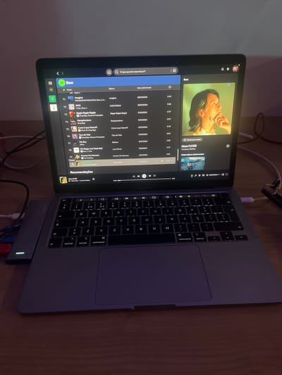 The scene, located in Pedrouços, Portugal, depicts an open, silver-colored Apple MacBook laptop resting on a light brown wooden surface. The laptop screen is illuminated, displaying a dark-themed music streaming application interface. On the left side of the screen, a playlist titled "Best" is visible, listing tracks with titles such as "Imagina," "DIAS," and "Biggie Biggie Biggie," along with their respective artists, albums, and added dates, predominantly from early 2024. The track "How I'd Kill" by "Cowboy Malley" is currently selected and playing, indicated by a highlighted entry and a progress bar at the bottom of the screen. The right side of the screen displays a prominent album art or video thumbnail featuring an individual with light brown hair, depicted in profile, looking upwards and to the right, with their right hand near their chin. This image is rendered with a warm, yellowish-green tint. The laptop's QWERTY keyboard is backlit and includes Portuguese-specific characters. A grey multi-port USB-C hub with a glowing white indicator light is connected to the laptop's left side, with red, blue, and white cables plugged into its ports. A white cable is also connected to the laptop's right side, extending off-frame. The immediate environment is dimly lit, with the laptop screen serving as the primary light source, casting subtle reflections on the wooden surface.