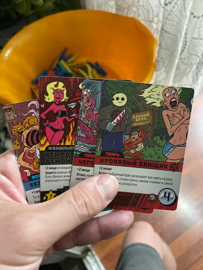 The image shows a person holding four cards from a card game. The cards have cartoonish illustrations and text in Russian. One card depicts a red-skinned devil woman with blonde hair holding a martini glass. Another card features a character in a hockey mask holding a bloody knife, standing over a smaller character. A third card shows a zombie-like creature. The fourth card has a yellow star symbol and a numeral 2. The cards are held in front of a yellow bowl and white lace fabric, with some green foliage visible in the background. The scene appears to be indoors. There is no information in the image to indicate the location is Novocherkassk, Russia.