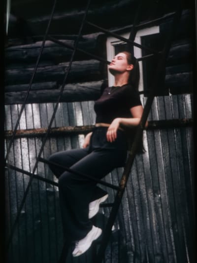 A young woman with brown hair pulled back in a ponytail is sitting on a metal ladder. She is wearing a dark t-shirt and dark pants, with white sneakers on her feet. Her head is tilted back, and her eyes are closed, giving the impression she is enjoying the moment. The ladder appears to be attached to a building with dark wooden siding. Behind the wooden siding is a corrugated metal wall. A white-framed window is visible above and to the right of the woman. The overall scene appears to be outdoors, possibly in a rural or industrial setting. The lighting suggests it could be daytime, though the dark tones of the building and surrounding elements create a somewhat somber atmosphere. There is no visible text in the image.