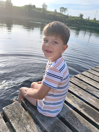 A young boy is sitting on a wooden dock facing the water. He is wearing a striped t-shirt with orange and navy blue stripes on a white background and dark blue shorts. The boy has short brown hair and is smiling at the camera. The water is calm with ripples and reflects the sky. In the background, there is a treeline and a clear blue sky with some clouds. The scene appears to be set in a natural environment, possibly a lake or river, near Brocēni, Latvia.