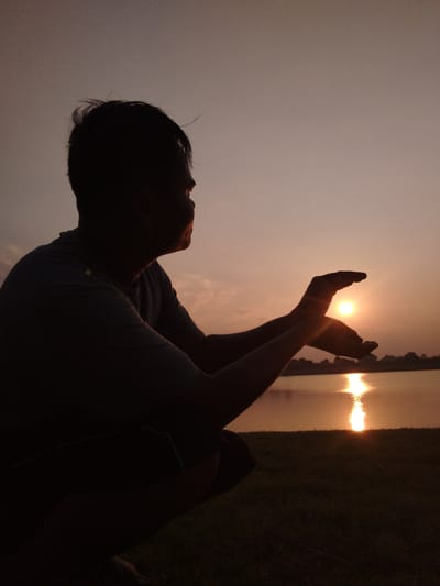 This is a silhouette of a person crouching near a body of water during sunset. The person is positioned in the foreground, with their back to the camera, looking out towards the setting sun. Their hands are held up in front of them, cupping the sun as if to hold it. The sun appears as a bright, warm orb low on the horizon, casting a shimmering reflection on the water. The sky is a soft orange and pink hue, characteristic of a sunset.

In the background, there is a calm body of water, likely a lake or river, with a faint outline of trees and buildings on the far bank. The ground in the foreground is covered in grass. The overall mood of the image is peaceful and contemplative, with a sense of awe and connection to nature. The time of day is clearly sunset, and the weather appears calm with clear skies. The location cues suggest an outdoor setting near a waterfront. There is no visible text in the image.