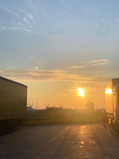 The image depicts a sunset at what appears to be a logistics or industrial area in Niepołomice, Poland. The sky is a gradient of pale blue and orange, with wispy clouds. The sun, a bright, elongated orb, is low on the horizon, casting a warm glow and creating reflections on the wet pavement in the foreground. Two large trucks are visible, one yellow on the left and a white one on the right, parked side-by-side. Their trailers are partially cut off by the frame. In the background, there are industrial structures, including fences with Y-shaped posts, streetlights, and possibly buildings. The ground is paved with interlocking bricks, and there are puddles of water. No people are visible in the scene.