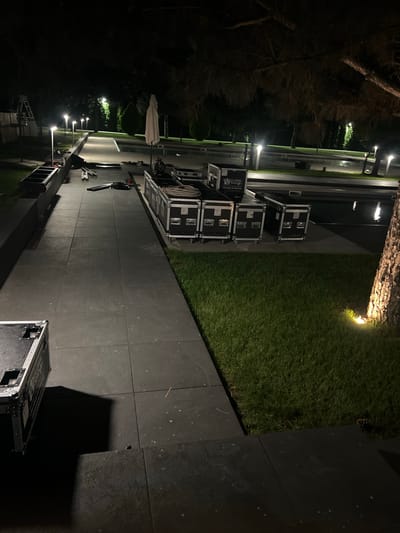 The image depicts an outdoor scene at night, likely in Vyshen'ky, Ukraine, where equipment cases are arranged along a paved pathway. The pathway, composed of dark square tiles, runs alongside a grassy area. Several large, black equipment cases with silver hardware are stacked and lined up. Some of these cases have branding visible, including "GLOBAL SHOW" and a phone number. In the background, a swimming pool is visible, reflecting the light from nearby lamps. A white umbrella stands near the pool, and trees and foliage surround the area, with some trees casting shadows. Several tall, illuminated lampposts are spaced along the pathway. The scene suggests preparations or cleanup for an event.