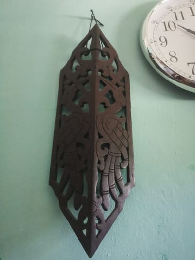 A dark, intricately carved wooden ornament, possibly a wall hanging, is suspended against a pale green wall. The ornament is elongated and features symmetrical designs that appear to depict stylized birds or mythical creatures with feathered tails. A portion of a round wall clock with a white face and black numbers is visible in the upper right quadrant of the image. The clock face shows the time as approximately 10:08. The environment appears to be indoors, possibly a residential or public space, in Cotraya, Indonesia. There are no people visible in the image.