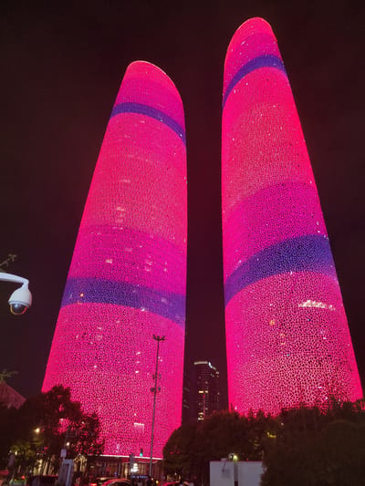 The image captures a nighttime view of two illuminated skyscrapers in the Wuhou District of China, likely Guangzhou. The buildings are predominantly covered in a vibrant pink light, with distinct purple bands circling each structure at different heights. The facades of the skyscrapers feature a intricate geometric pattern of lights, creating a textured effect. In the foreground, a security camera is visible mounted on a white pole. The scene is set against a dark night sky, with the lights of other buildings and streetlights visible in the distance. Trees and foliage are present at the base of the skyscrapers, and there are hints of traffic and pedestrians on the street level.