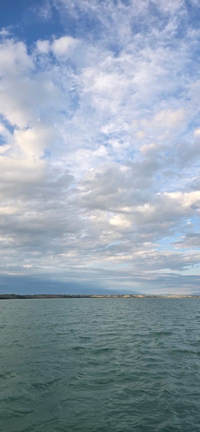The image depicts a wide expanse of choppy, teal-colored water in the foreground, reflecting the sky above. The horizon line is visible, separating the water from a distant, flat shoreline. Beyond the shoreline, a range of muted, mountainous terrain stretches across the background, partly obscured by atmospheric haze. The sky is filled with a variety of clouds, ranging from scattered, wispy white clouds higher up to denser, grayer formations closer to the horizon. The lighting suggests it may be late afternoon or early evening, with a soft, diffused light illuminating the scene. The overall impression is one of a serene natural landscape, likely a lake or a large body of water. The city of Pristan'-Przheval'sk, Kyrgyzstan, is situated on the eastern shore of Lake Issyk-Kul, which aligns with this visual description.