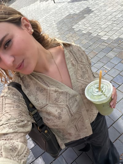 A woman is taking a selfie outdoors, in a location resembling the city of Coral Gables, United States of America. She is holding a plastic cup with a green-colored drink and a straw, positioned in front of her. She is wearing a beige, patterned, button-up shirt, dark pants, and a black shoulder bag. She has light-colored hair and is wearing earrings. The background shows a brick or stone-paved area, suggesting an urban setting.
