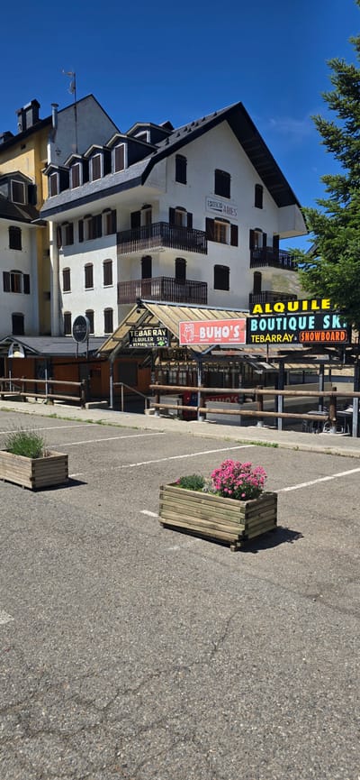 The image shows a view of a building in Sallent de Gállego, Spain, with businesses on the ground floor. The building is white with dark shutters and balconies. Above the ground floor businesses, there is a sign for "EDIFICIO ABIES". The businesses include "TEBARRAY ALQUILER SKI REPARACION", "BUHO'S", and "ALQUILER BOUTIQUE SKI TEBARRRAY SNOWBOARD". There are wooden planters with greenery and pink flowers in the foreground, placed in a paved parking lot. The sky is clear and blue.