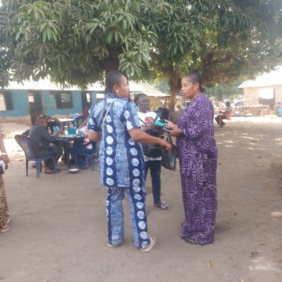 The image captures an outdoor scene in Assai Sabongida, Nigeria, under natural daylight. In the foreground, three adult women stand on unpaved, light-brown ground. On the left, a woman in a blue and white patterned two-piece outfit (tunic and trousers) and light flip-flops holds a black bag in her left hand and extends her right hand towards a woman standing to her right. This second woman, wearing a long purple patterned dress, holds a blue-green electronic device with both hands, facing the first woman. A third woman stands slightly behind the interaction, dressed in a white t-shirt with a red logo and dark trousers, observing.
Large, leafy trees dominate the upper portion of the image, providing overhead shade. In the midground to the left, several individuals are seated at plastic tables on plastic chairs, engaged in activities such as eating or conversing; a man in a dark shirt is clearly visible among them. In the background, simple buildings with corrugated metal roofs are visible, including one structure with blue-painted walls. The environment appears to be an open public area, possibly a community gathering spot or a casual market setting.