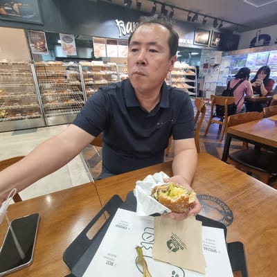 The image depicts an indoor scene at a bakery in Wonju-si, South Korea. A middle-aged East Asian man is seated at a wooden table, holding a half-eaten burger in his right hand. He is wearing a dark blue polo shirt with a small white logo on the left chest. In front of him on the table is a black phone, a white paper wrapper with burger branding, and a menu or placemat. To his right, a circular graphic with the text "NEW YORK BURGER" is visible on the table surface. In the background, shelves filled with baked goods are displayed, and other patrons are seated at tables. One woman with glasses and another with a backpack are visible in the background, along with chairs and tables. The overall environment suggests a casual dining or bakery setting.