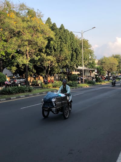 The media file captures a street scene in Gelam, East Java. The primary subject is a person riding a three-wheeled cart pulled by a motorcycle. The cart is loaded with various items, including what appears to be blue tarpaulin, white bags, and possibly other belongings. The rider is wearing a light blue shirt and a dark cap.

The scene is set on a paved road with a clear lane divider. A sidewalk with green vegetation and trees lines the road. In the background, more trees, buildings, and other vehicles, including motorcycles and a black car, are visible. The time of day appears to be late afternoon or early evening, as indicated by the soft lighting and shadows. The weather seems pleasant, with a clear sky and some clouds.

The overall impression is of a bustling street, likely in a semi-urban or suburban area, with daily life in progress. There is no visible text in the image. The activity depicts transportation and perhaps the movement of goods or personal effects. The rider's posture suggests they are actively engaged in operating the vehicle and managing the load. The presence of other vehicles and pedestrians implies a public thoroughfare.