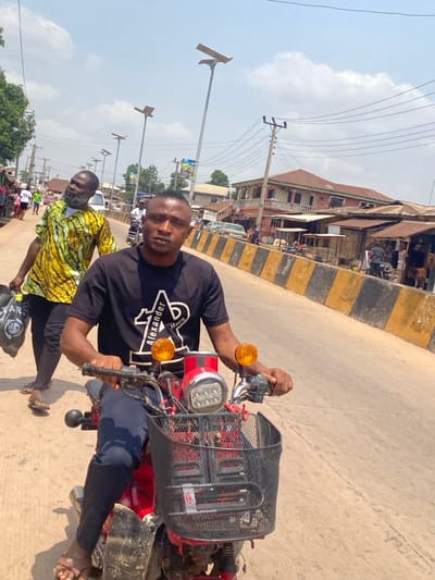 This image depicts a daytime street scene in the city of Afikpo, Nigeria. In the foreground, a dark-skinned male individual, appearing to be in his 20s or 30s, is seated on a red motorcycle, looking directly at the camera. He wears a black t-shirt with "Alexander Macs" branding and dark trousers, with brown sandals on his feet. The motorcycle features a front-mounted black metal basket, a central chrome headlight with multiple LED lights, and two prominent orange turn indicators. His hands rest on the handlebars.
Immediately behind and to the left of the motorcyclist, another dark-skinned male, older with a beard, walks towards the left, carrying a black plastic bag in his left hand. He is dressed in a yellow and green patterned short-sleeved shirt and dark trousers, and is also wearing sandals. His head is tilted upwards and to his left.
The environment includes a paved road surface with a distinct yellow and black striped median or curb running along the right side. On both sides of the road, various buildings are visible, ranging from multi-story structures with balconies to simpler stalls with corrugated metal roofs, suggesting commercial activity. Numerous utility poles with power lines traverse the scene, accompanied by modern, tall streetlights equipped with solar panels. Distant vehicles and pedestrians indicate active street traffic under a bright, partly cloudy sky.