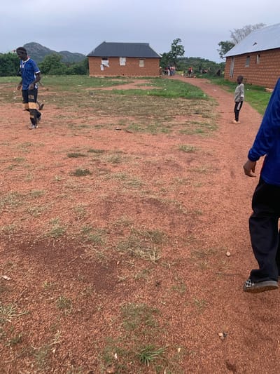 In the city of Buga da Gindi, Nigeria, a group of individuals, primarily young boys, are engaged in an activity that appears to be a casual soccer game on a dirt field. The field itself is a reddish-brown color, sparsely covered with dry grass and scattered with small stones and debris. Two houses constructed from mud bricks with corrugated metal roofs are visible in the background, suggesting a rural or semi-urban setting.

In the foreground, a young boy in a light gray long-sleeved shirt and black pants with white stripes stands facing forward, with a slight smile. To his left, a young man in a blue soccer jersey with black and gold shorts is mid-stride, appearing to be running or playing soccer. He has a headband on and a determined expression. His soccer cleats are visible. Further back, other figures, including more children and possibly adults, are scattered around the scene, some observing, others participating in the activity. The environment is characterized by an overcast sky, with a hilly landscape in the far distance. The overall impression is one of communal activity and everyday life in a Nigerian village.