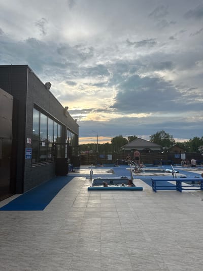 In Yuzhnyye Koryaki, Russia, an outdoor recreational facility features a pool area with several small, tiled pools, some with blue decking and benches. Several individuals are present in and around the pools, some bathing and others standing or sitting. A dark-colored building with large windows, possibly a changing room or cafe, is situated to the left of the scene, with a blue mat extending from its entrance towards the pool area. The sky above is overcast with dramatic clouds, and the sun is setting, casting a warm glow on the horizon. The overall environment appears to be a public leisure space.