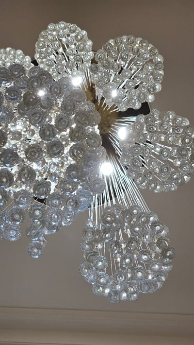 This media file shows a close-up, low-angle view of a crystal chandelier. The chandelier is a prominent object in the scene, with numerous spherical, faceted crystal elements arranged in clusters and suspended by thin wires. The crystals catch and refract light, creating a glittering effect. Several light bulbs are visible within the chandelier, illuminating the crystals from within and contributing to the bright, sparkling appearance. The background is out of focus and appears to be a wall with a neutral, light-colored tone, possibly suggesting an indoor setting like a room or hallway. The lighting suggests it could be daytime or evening with artificial lights on, as the chandelier is the primary light source in the frame. There is no discernible activity taking place, as the focus is solely on the static object. No people are visible. There is no visible text in the image. The context provided, Kamenka, Russia, does not offer specific visual cues within the image itself.